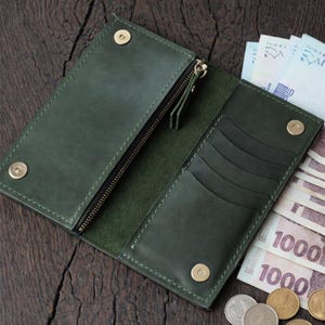 May include: Open, dark green leather wallet with a zipper compartment and multiple card slots. The wallet is secured with snap closures. Several bills and coins are visible next to the open wallet.
