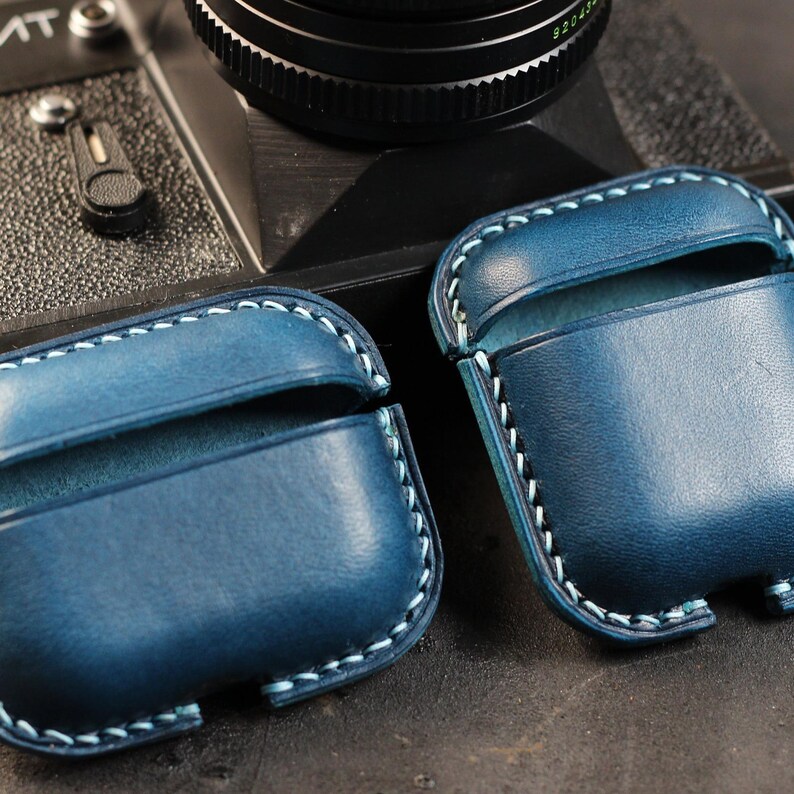 May include: Two blue leather cases with white stitching for AirPods. The cases are open and show the interior lining.