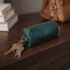 May include: A dark teal leather key pouch with a brass zip and a small tassel. Several keys are attached to a brass ring on the pouch. The pouch is resting on a dark wooden surface.