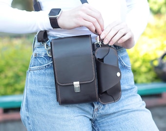 May include: A dark brown leather crossbody bag with a matching sunglasses case attached. The bag has a rectangular shape with a flap closure and silver hardware. The sunglasses case is secured with a button. The bag is worn with a brown strap.