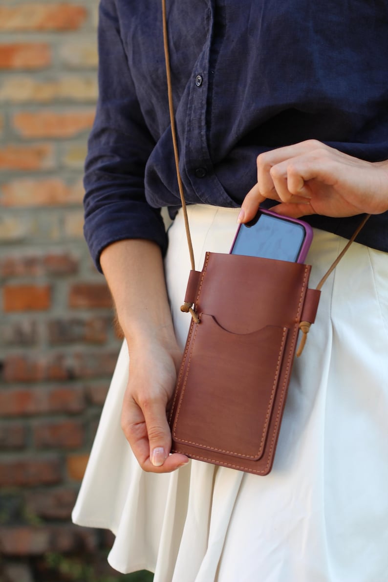 Op de afbeelding: Een bruine leren crossbody telefoonhoes met een lange riem. De hoes heeft een zak aan de voorkant en een hoofdvak. De telefoon is paars en zwart. De hoes wordt door een persoon vastgehouden.