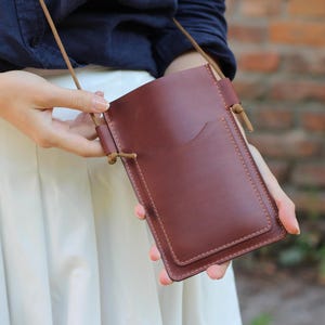 May include: A brown leather crossbody phone bag with a flap closure and a small pocket on the front. The bag has a brown leather strap.