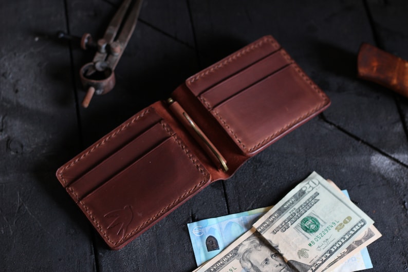 May include: A brown leather wallet with a money clip and multiple card slots. The wallet is open and has cash inside.