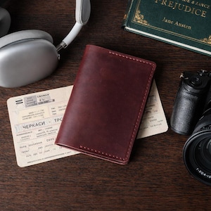 May include: A burgundy leather passport holder rests on a document, next to a pair of white headphones, a camera, and a book titled "Pride & Prejudice" by Jane Austen. The passport holder is rectangular and appears to be hand-stitched.