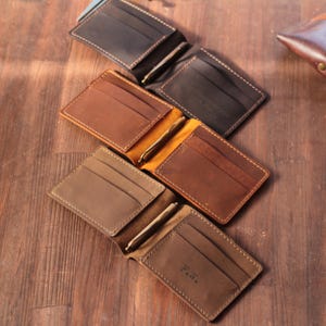 Puede incluir: Cuatro carteras de cuero abiertas en varios tonos de marrón, que muestran ranuras para tarjetas y pinzas para billetes. Las carteras están dispuestas sobre una superficie de madera, con costuras visibles y una inicial personalizada en una cartera.