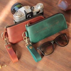 Könnte beinhalten: Zwei Leder-Reißverschlusstaschen, eine braune und eine grüne, mit Handschlaufen und eine schwarze Sonnenbrille. Im Hintergrund eine Vintage-Kamera. Auf der grünen Tasche steht "I love I need I want".