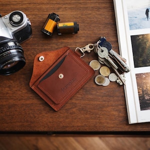 May include: A brown leather wallet with the name "Stanislav" embossed on the front, open to reveal a black interior. A vintage camera, film rolls, keys, and coins are arranged on a wooden surface.