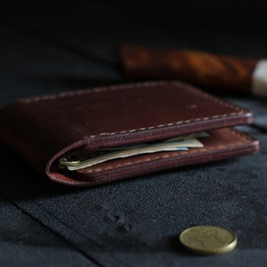 May include: A brown leather wallet with a stitched edge, folded closed and lying on a dark wooden surface. The wallet is filled with cash and has a worn look.