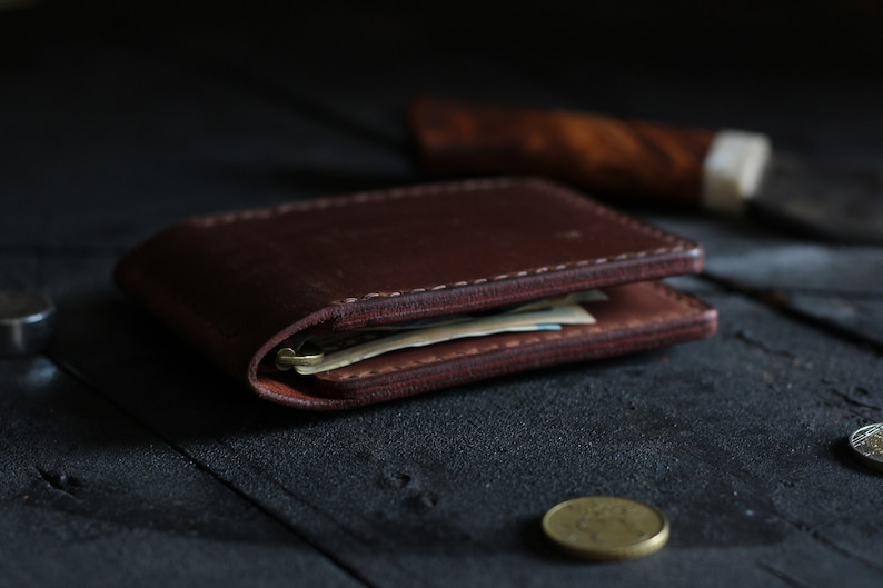 May include: A brown leather wallet with a stitched edge, folded closed and lying on a dark wooden surface. The wallet is filled with cash and has a worn look.