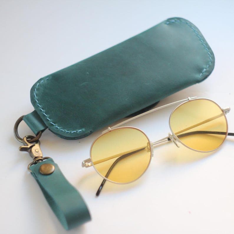 May include: A teal leather glasses case with a matching wrist strap and gold-tone hardware. The case is next to a pair of round, yellow-tinted sunglasses with a silver frame.