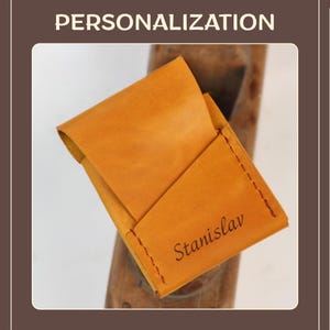 May include: A handcrafted, open leather cardholder in a warm, golden-brown hue. The cardholder features a stitched edge with red thread and the name "Stanislav" engraved on the front. The word "PERSONALIZATION" is at the top.