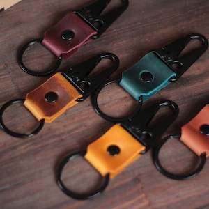 Könnte beinhalten: Fünf Leder-Schlüsselanhänger mit schwarzen Metallhaken und Ringen. Die Schlüsselanhänger sind in verschiedenen Farben: Burgunderrot, Braun, Türkis, Orange und Beige.