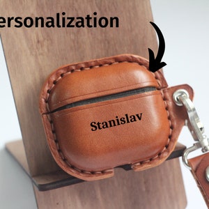 May include: A brown leather Airpods case with a personalized name "Stanislav" engraved on the front. The case has a stitched edge and a silver metal hook with a brown leather strap.