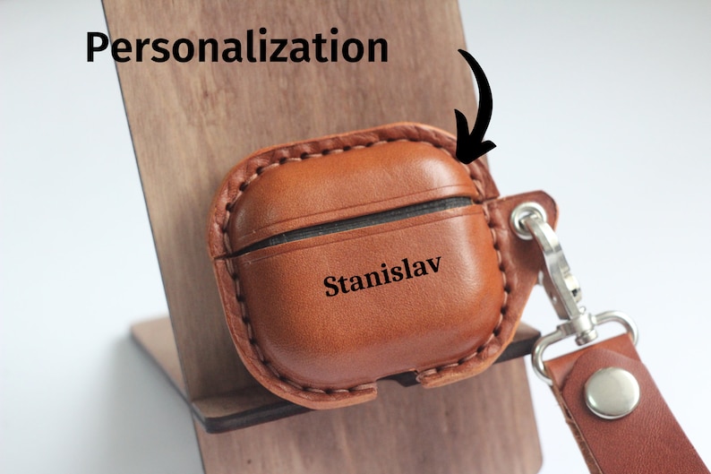 May include: A brown leather Airpods case with a personalized name "Stanislav" engraved on the front. The case has a stitched edge and a silver metal hook with a brown leather strap.