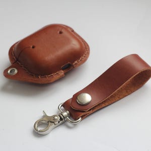 May include: A brown leather case for AirPods with a matching brown leather strap and a silver metal carabiner clip.