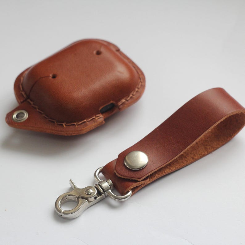 May include: A brown leather case for AirPods with a matching brown leather strap and a silver metal carabiner clip.