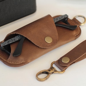 May include: A brown leather glasses case with a pair of black-framed glasses inside. A matching leather keychain with a brass clasp and button is next to the case. The case is closed with a brass button.