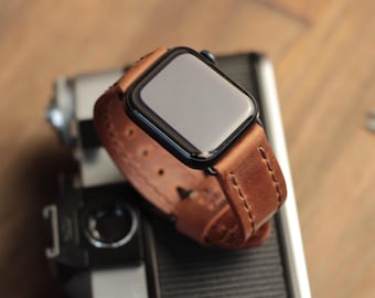 May include: A brown leather watch band with white stitching, attached to a black Apple Watch.
