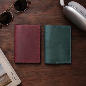 Può includere: Due porta passaporto rettangolari in pelle, uno bordeaux scuro e uno verde bosco, con cuciture visibili lungo i bordi. Gli oggetti sono posizionati accanto a un paio di occhiali da sole con montatura dorata e cuffie wireless argentate.