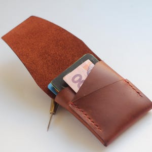 May include: A brown leather cardholder with red stitching, open to reveal cards and cash. The wallet is made of a rich, textured leather. A small, gold-colored tool is next to the wallet.