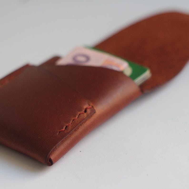 May include: A brown leather wallet with a folded design, showcasing a glimpse of the interior. The wallet is filled with folded paper money. The edges are stitched with red thread, adding a contrasting detail.