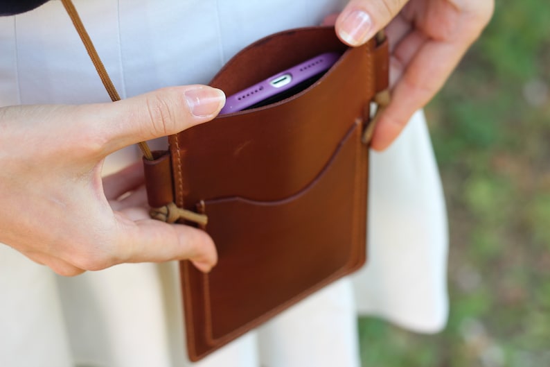 Puede incluir: Una funda de tel&eacute;fono de cuero marr&oacute;n con una correa. La funda tiene una solapa que se cierra con un broche. Un tel&eacute;fono morado est&aacute; dentro de la funda.