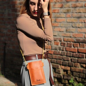 Op de afbeelding: Een bruinleren telefoon crossbody tas met een gele riem. De tas heeft een zak aan de voorkant en de naam "Sue" is op de onderkant gestempeld. De tas wordt gedragen over een grijze broek en een bruine riem.