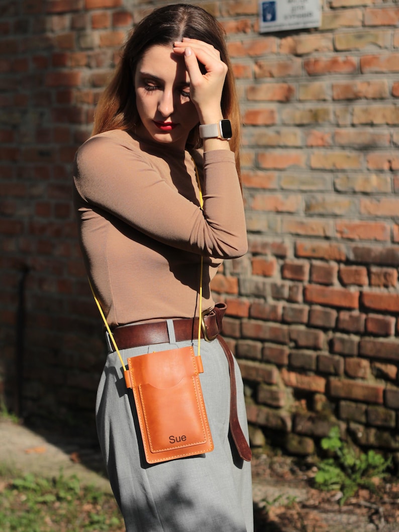 Op de afbeelding: Een bruinleren telefoon crossbody tas met een gele riem. De tas heeft een zak aan de voorkant en de naam "Sue" is op de onderkant gestempeld. De tas wordt gedragen over een grijze broek en een bruine riem.