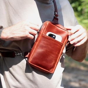 May include: A brown leather crossbody bag with a front pocket and a zip closure. The bag has a strap that goes over the shoulder and a belt that goes around the waist. A white smartphone is visible in the front pocket.