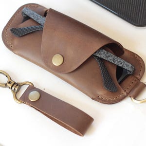 May include: A brown leather glasses case with a snap closure, holding a pair of black-framed glasses. A matching leather keychain with a brass clasp is next to the case. The case and keychain are made of brown leather.