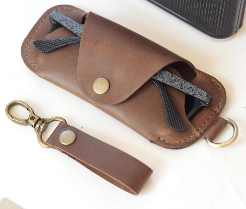 May include: A brown leather glasses case with a snap closure, holding a pair of black-framed glasses. A matching leather keychain with a brass clasp is next to the case. The case and keychain are made of brown leather.