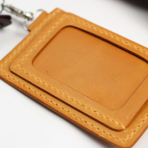 May include: A tan leather ID holder with a rectangular window, stitched edges, and a silver-colored clasp. The holder is designed to display an ID card and is attached to a keyring with keys.