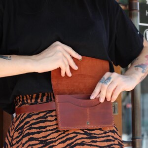 May include: A brown leather belt bag with a flap closure and multiple compartments. The bag is worn around the waist and has a matching brown leather belt. The bag is open, revealing the interior. The person is wearing a black top and a skirt with an orange and black pattern.