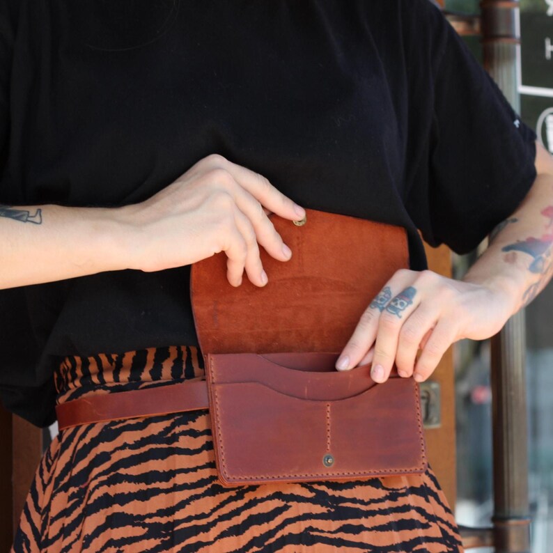 May include: A brown leather belt bag with a flap closure and multiple compartments. The bag is worn around the waist and has a matching brown leather belt. The bag is open, revealing the interior. The person is wearing a black top and a skirt with an orange and black pattern.