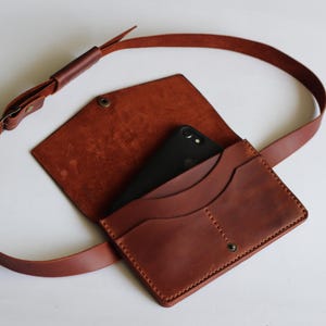 May include: A brown leather crossbody bag with a flap and long strap. The bag has a front pocket and a snap closure. A black smartphone is visible inside. The strap is adjustable and made of the same brown leather. The bag is suitable for everyday use.