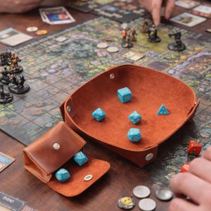May include: A brown leather dice tray and pouch set with blue dice. The dice tray is square with rounded corners and a snap closure. The dice are light blue with white markings. The set is on a game board.