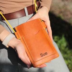 Op de afbeelding: Een bruinleren telefoonhoesje met een gele riem. De hoes heeft een voorvak en de naam "Sue" staat op de voorkant. De hoes wordt door een persoon vastgehouden.