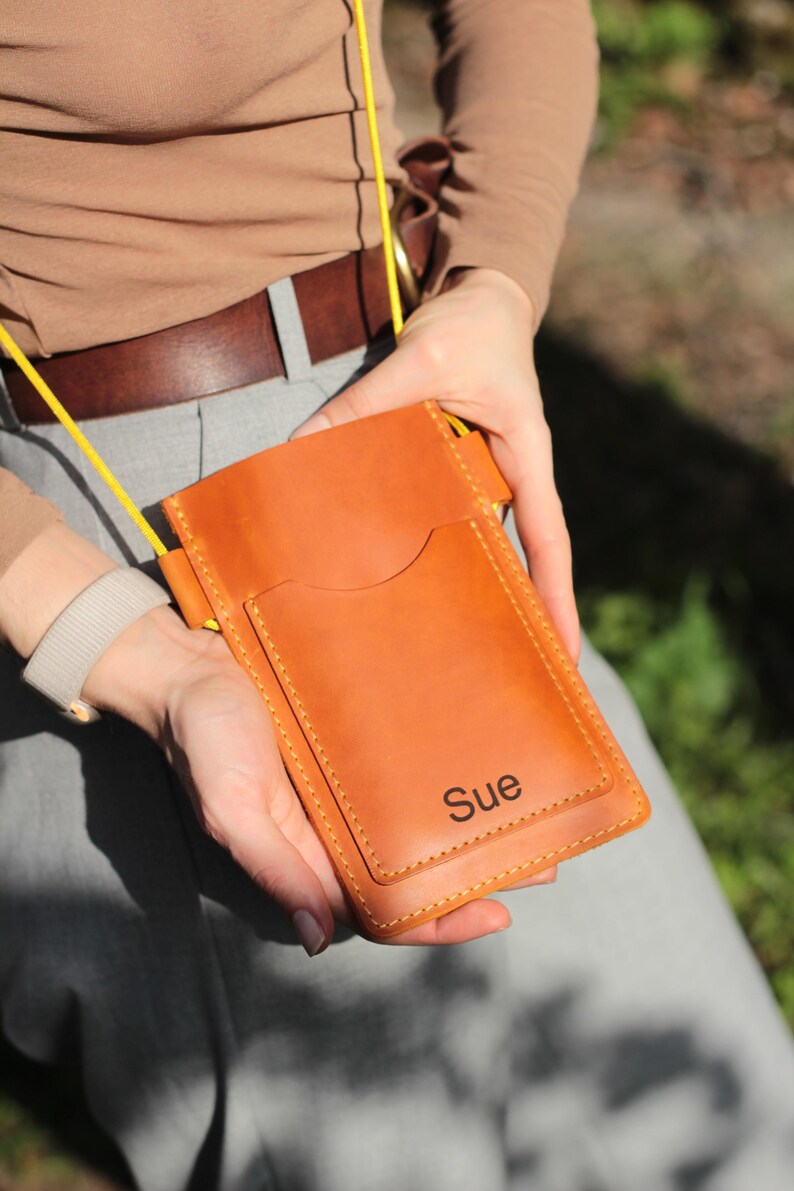 Op de afbeelding: Een bruinleren telefoonhoesje met een gele riem. De hoes heeft een voorvak en de naam "Sue" staat op de voorkant. De hoes wordt door een persoon vastgehouden.