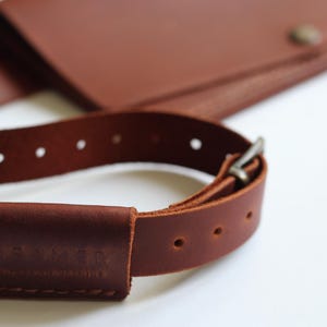 May include: Close-up of a brown leather bag with a matching strap. The strap has a buckle and several holes for adjustment. The bag and strap are made of high-quality leather.