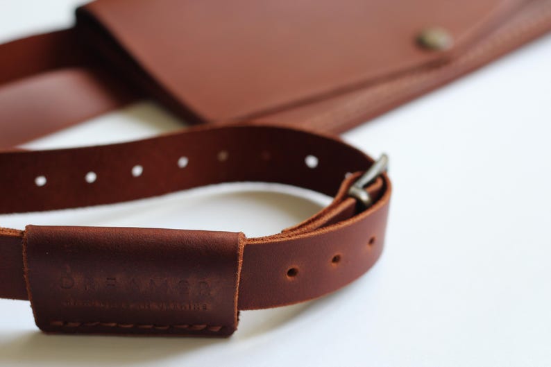 May include: Close-up of a brown leather bag with a matching strap. The strap has a buckle and several holes for adjustment. The bag and strap are made of high-quality leather.