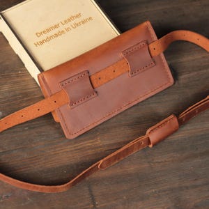 May include: A brown leather fanny pack with an adjustable strap and buckle. The pack is rectangular with stitched detailing and a leather belt loop. The box next to it reads "Dreamer Leather Handmade in Ukraine."