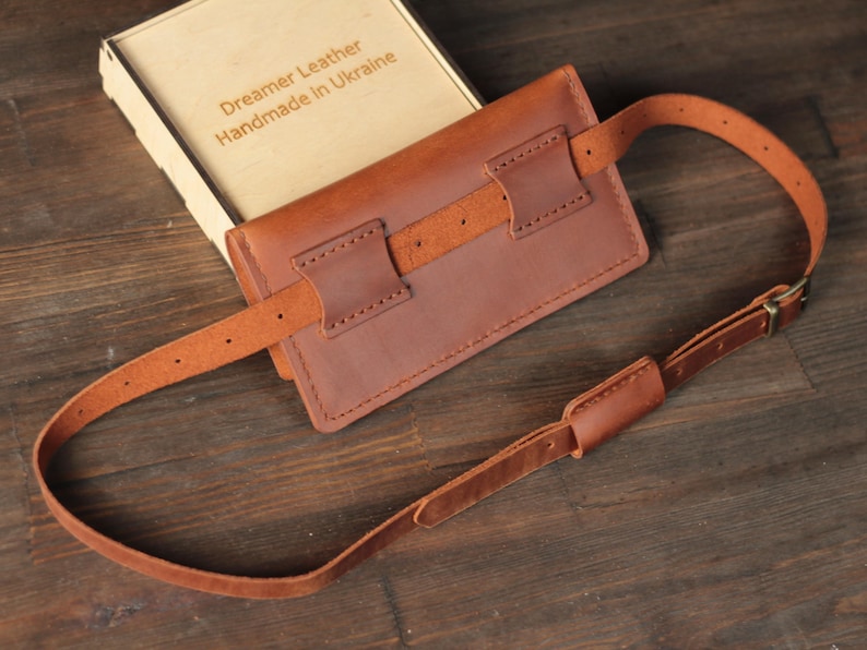 May include: A brown leather fanny pack with an adjustable strap and buckle. The pack is rectangular with stitched detailing and a leather belt loop. The box next to it reads "Dreamer Leather Handmade in Ukraine."