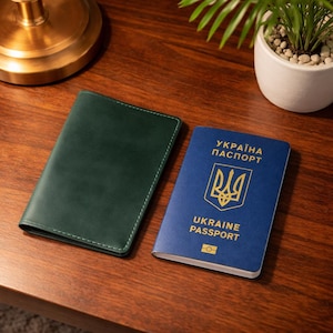 May include: A dark green leather passport holder next to a blue Ukraine passport. The passport has gold lettering and a gold emblem. The passport holder is rectangular with white stitching around the edges. The items are on a dark wood surface.