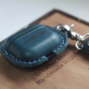 May include: A blue leather Airpods case with a light blue stitching detail. The case has the words "soul therapy" engraved on the front. The case is handmade in the UK and is made by Dreamer Leather.