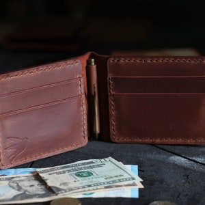 May include: A brown leather wallet with a money clip and multiple card slots. The wallet is open and has cash and cards inside.
