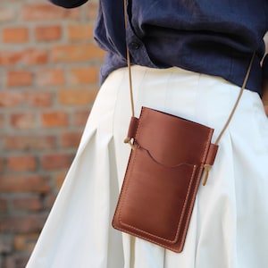 May include: A brown leather phone case with a flap closure and a small pocket on the front. The case is attached to a long brown strap and is worn over the shoulder.