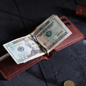 May include: A brown leather wallet with a money clip holding several US twenty dollar bills. The wallet has a stitched edge and the text "monobank" is visible on the inside.