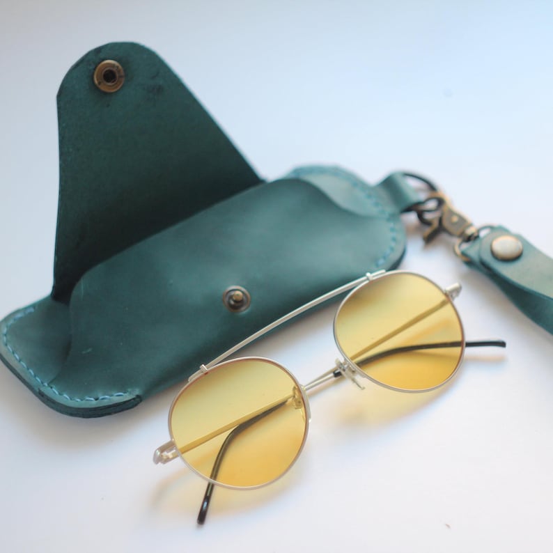 May include: A teal leather case with a snap closure and a pair of round, yellow-tinted sunglasses with a silver frame.