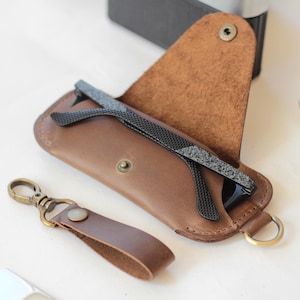 May include: A brown leather sunglasses case with a pair of sunglasses inside. A matching leather keychain with a brass clasp is next to the case. The case is open, revealing the sunglasses.