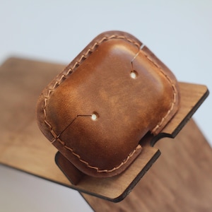 May include: A brown leather case with stitching around the edges. The case has two small holes in the top. The case is sitting on a wooden stand.